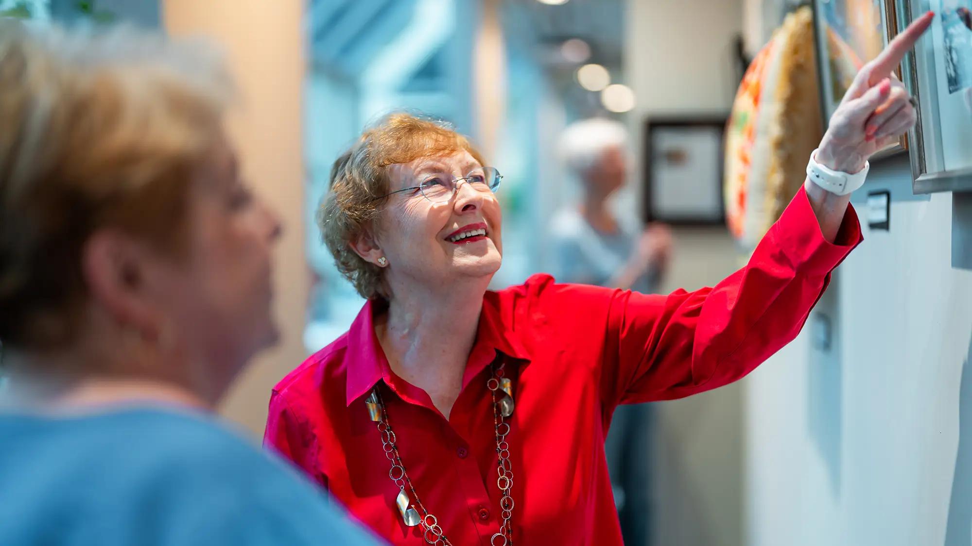 Woman in red shirt pointing at art on the wall while talking to woman in blue shirt.