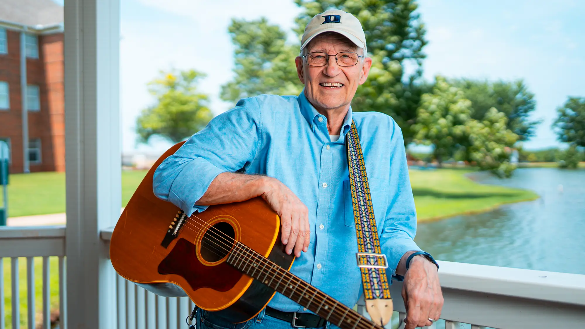 Man in hat and blue button down shirt holding an acoustic guitar.