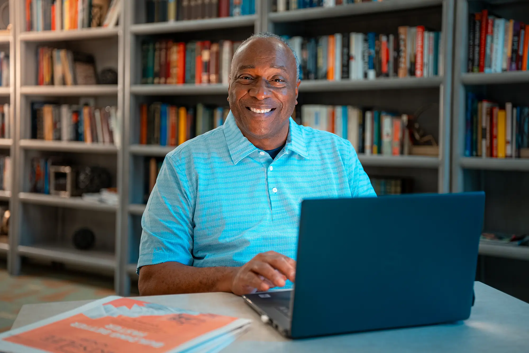 Man in teal polo shirt sitting in front of a bookshelf with a laptop open