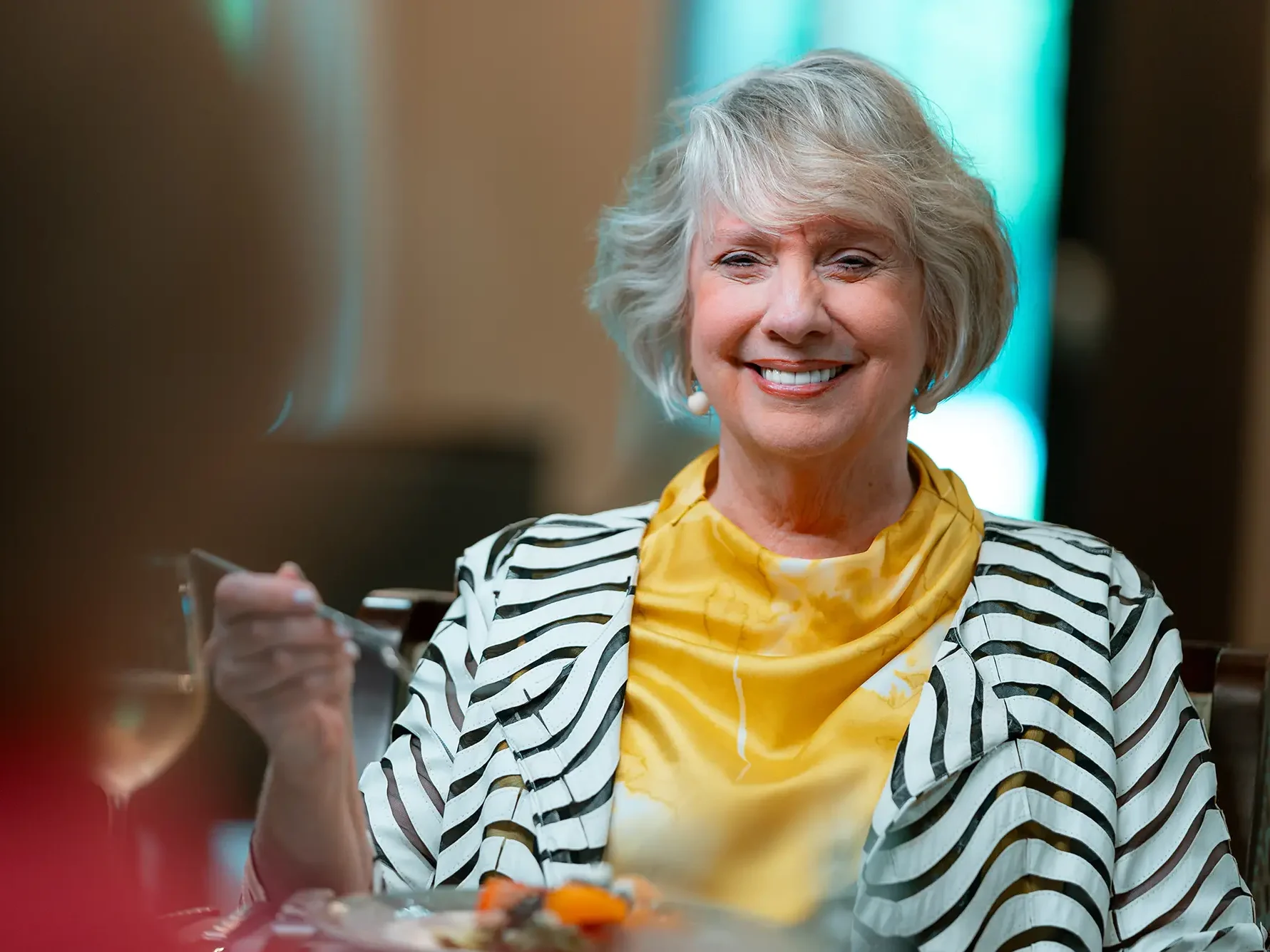 Woman in yellow shirt with striped jacket smiling holding a fork.